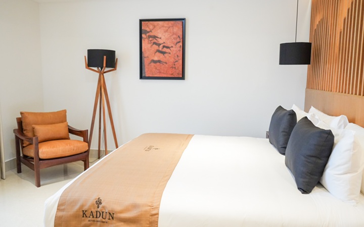 Luxury suite interior showcasing a comfortable King-size bed, a plush accent lounge chair, and a sophisticated lighting design with a hanging ceiling lamp and a modern pedestal floor lamp at Kadun Hotel Boutique.